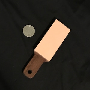 Puede incluir: Un asentador de cuero con un mango de madera y un agujero para colgar. El asentador tiene una superficie rosa claro y un mango marrón oscuro. Una moneda de plata está al lado del asentador.