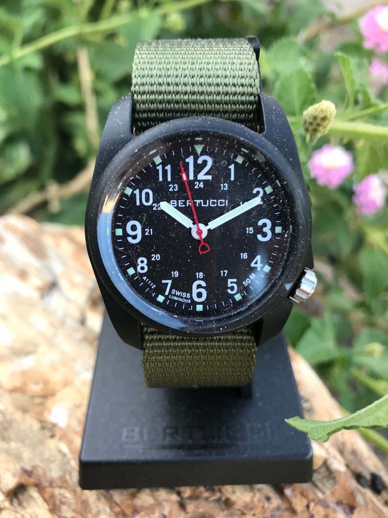 May include: A black and green military style watch with a black face and white numbers. The watch face also has the text "BERTUCCI" and "50M" printed on it. The watch is on a black stand.