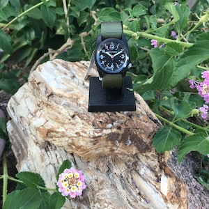 May include: A black wristwatch with a green nylon strap. The watch face has white numbers and a red second hand. The watch is on a black stand and is surrounded by green foliage.