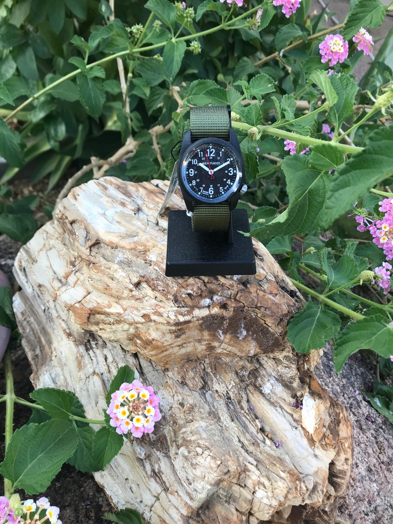 May include: A black wristwatch with a green nylon strap. The watch face has white numbers and a red second hand. The watch is on a black stand and is surrounded by green foliage.