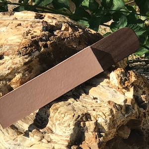 Puede incluir: Un marcapáginas de cuero marrón con un extremo de madera marrón oscuro. El marcapáginas está apoyado en un trozo de madera petrificada.