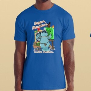 May include: Royal blue t-shirt with a cartoon manatee artist wearing glasses and a beret, painting on a canvas. The shirt features the text "Dapper Manatees" and "Custom Creations" in white script.