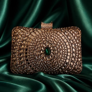 May include: A rectangular gold-toned clutch bag featuring an intricate pattern of embedded clear crystals and small mirrors. A single oval-shaped emerald green gemstone is centered on the front surface, and a metallic clasp sits at the top.