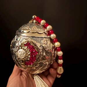 May include: A spherical silver-toned metal clutch bag featuring intricate embossed floral patterns, a central circular medallion with red crystals and a white faux pearl, a beaded handle of red and white beads, and a white beaded tassel hanging from the base.