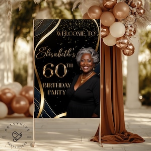 May include: A welcome sign for a 60th birthday party. The sign features a photo of a smiling woman with gray hair, and the text "Welcome to Elisabeth's 60th Birthday Party" in gold lettering against a black background with gold accents. The sign is displayed on a gold stand.