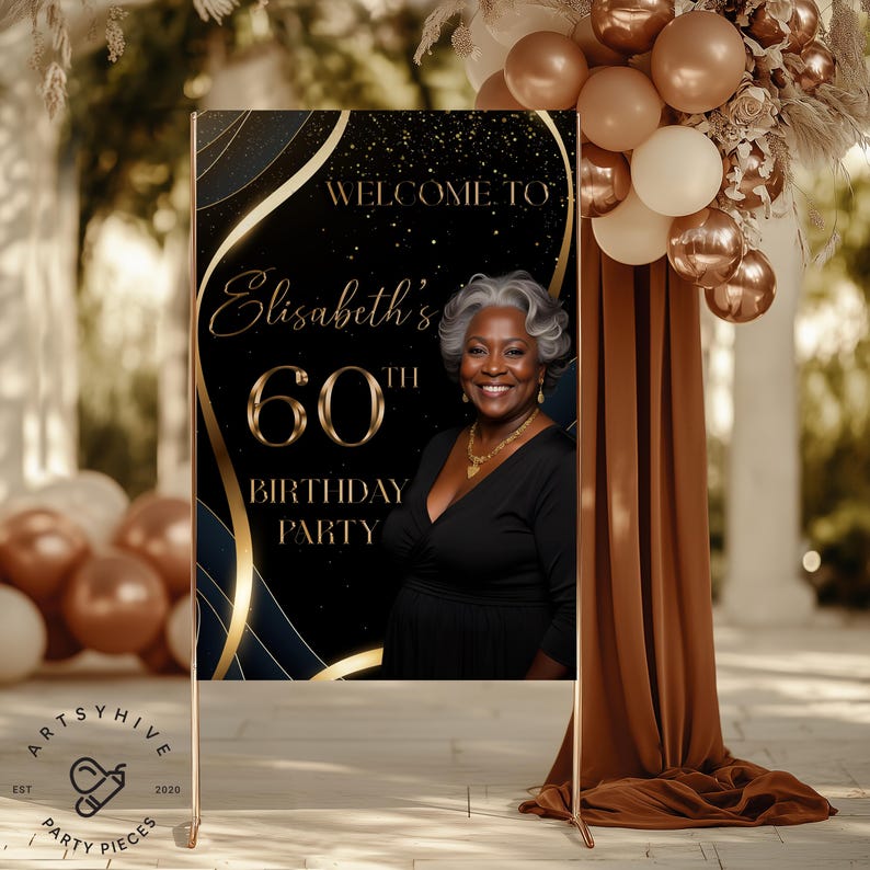 May include: A welcome sign for a 60th birthday party. The sign features a photo of a smiling woman with gray hair, and the text "Welcome to Elisabeth's 60th Birthday Party" in gold lettering against a black background with gold accents. The sign is displayed on a gold stand.