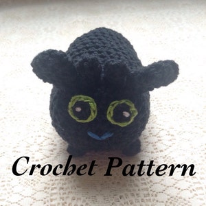 May include: Black crochet amigurumi toy with green eyes and a blue mouth. The toy is shaped like a dragon and has a textured surface. Crochet Pattern.