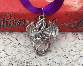 May include: Silver dragon pendant with wings outstretched, hanging from a purple ribbon. The pendant is on a white lace background.