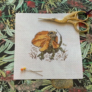 May include: A cross-stitch of a frilled-neck lizard in orange, yellow, and green. Gold scissors and pins are placed near the stitched design on white fabric. The background is a floral pattern with green leaves and red flowers.