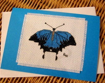 May include: A blue and black butterfly cross-stitch design on a white card with a blue border. The card is signed "RH".