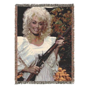 May include: A woven throw blanket featuring a portrait of a blonde individual with voluminous hair wearing a white ruffled top, holding a long-barreled firearm. The background displays autumn foliage with orange and yellow tones and fringed edges.