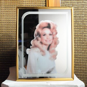 May include: A rectangular wall mirror with a thin gold frame featuring a centered, full-color portrait of a person with voluminous blonde hair and a white top. The mirror glass is etched with a decorative triple-line border in the upper left corner.