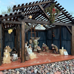 Puede incluir: Un bel&eacute;n de madera con figuras de Mar&iacute;a, Jos&eacute;, el ni&ntilde;o Jes&uacute;s, &aacute;ngeles y animales. La escena est&aacute; ambientada dentro de una estructura de madera con un techo de listones y paneles azules de fondo. Pacas de heno y velas rodean las figuras.