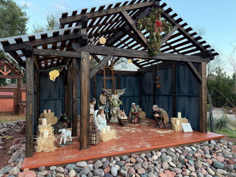 Puede incluir: Un bel&eacute;n de madera con figuras de Mar&iacute;a, Jos&eacute;, el ni&ntilde;o Jes&uacute;s, &aacute;ngeles y animales. La escena est&aacute; ambientada dentro de una estructura de madera con un techo de listones y paneles azules de fondo. Pacas de heno y velas rodean las figuras.