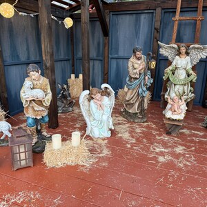 Puede incluir: Un bel&eacute;n navide&ntilde;o con varias estatuas de figuras religiosas, incluyendo a Mar&iacute;a, Jos&eacute;, &aacute;ngeles y el ni&ntilde;o Jes&uacute;s. Las figuras est&aacute;n dispuestas sobre un suelo de madera roja, con balas de heno y una linterna. El fondo presenta vigas de madera y paneles azules.
