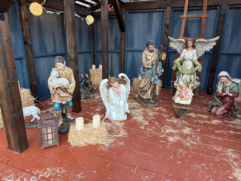 Puede incluir: Un bel&eacute;n navide&ntilde;o con varias estatuas de figuras religiosas, incluyendo a Mar&iacute;a, Jos&eacute;, &aacute;ngeles y el ni&ntilde;o Jes&uacute;s. Las figuras est&aacute;n dispuestas sobre un suelo de madera roja, con balas de heno y una linterna. El fondo presenta vigas de madera y paneles azules.