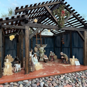 Puede incluir: Un bel&eacute;n de madera con figuras de Mar&iacute;a, Jos&eacute;, el ni&ntilde;o Jes&uacute;s, &aacute;ngeles y animales. La escena est&aacute; ambientada dentro de una estructura de madera con un techo de listones y un suelo marr&oacute;n rojizo. La estructura est&aacute; rodeada de rocas.