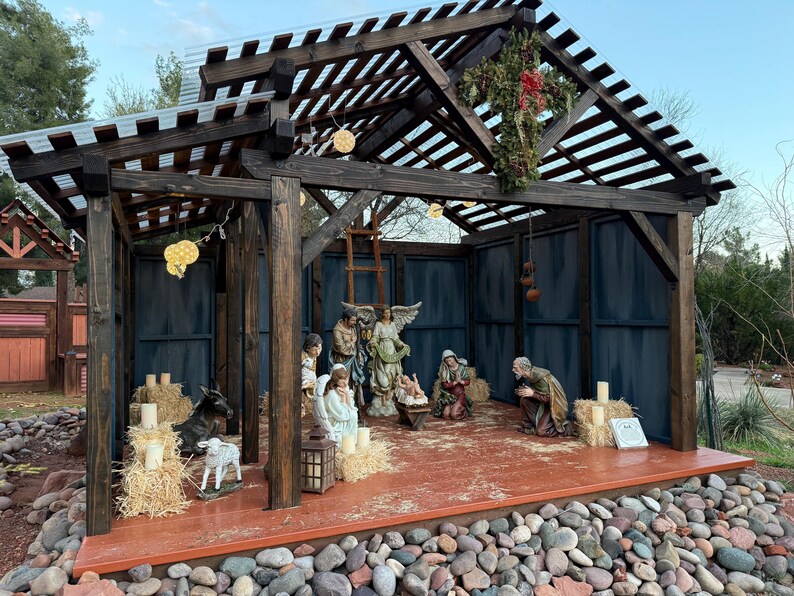 Puede incluir: Un bel&eacute;n de madera con figuras de Mar&iacute;a, Jos&eacute;, el ni&ntilde;o Jes&uacute;s, &aacute;ngeles y animales. La escena est&aacute; ambientada dentro de una estructura de madera con un techo de listones y un suelo marr&oacute;n rojizo. La estructura est&aacute; rodeada de rocas.