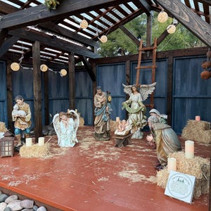 Puede incluir: Un bel&eacute;n navide&ntilde;o con varias estatuas de figuras b&iacute;blicas, incluyendo a Mar&iacute;a, Jos&eacute; y &aacute;ngeles. Las figuras est&aacute;n dispuestas en una estructura de madera con balas de heno y velas. La escena est&aacute; iluminada por luces de cadena.