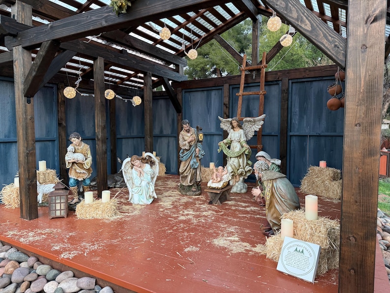Puede incluir: Un bel&eacute;n navide&ntilde;o con varias estatuas de figuras b&iacute;blicas, incluyendo a Mar&iacute;a, Jos&eacute; y &aacute;ngeles. Las figuras est&aacute;n dispuestas en una estructura de madera con balas de heno y velas. La escena est&aacute; iluminada por luces de cadena.