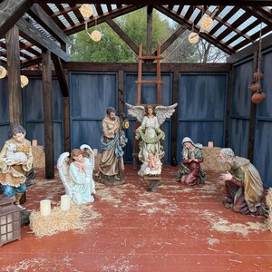 Puede incluir: Un bel&eacute;n con varias estatuas de figuras religiosas, incluyendo &aacute;ngeles, Mar&iacute;a, Jos&eacute; y un pastor. La escena est&aacute; ambientada dentro de una estructura de madera con luces de cadena y velas.