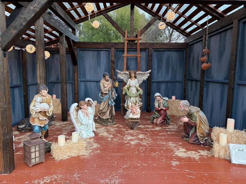 Puede incluir: Un bel&eacute;n con varias estatuas de figuras religiosas, incluyendo &aacute;ngeles, Mar&iacute;a, Jos&eacute; y un pastor. La escena est&aacute; ambientada dentro de una estructura de madera con luces de cadena y velas.