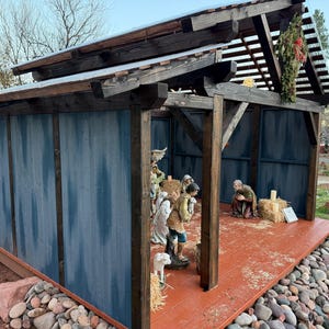 Puede incluir: Un bel&eacute;n de madera con un exterior pintado de azul y un suelo rojo-naranja. La estructura tiene un tejado inclinado y est&aacute; decorada con una peque&ntilde;a corona. Figuras del bel&eacute;n son visibles en el interior.