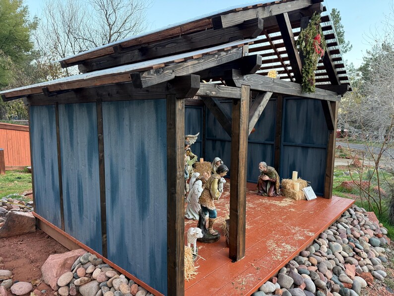 Puede incluir: Un bel&eacute;n de madera con un exterior pintado de azul y un suelo rojo-naranja. La estructura tiene un tejado inclinado y est&aacute; decorada con una peque&ntilde;a corona. Figuras del bel&eacute;n son visibles en el interior.