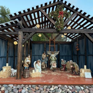 Puede incluir: Un bel&eacute;n navide&ntilde;o con figuras de Mar&iacute;a, Jos&eacute;, &aacute;ngeles y el ni&ntilde;o Jes&uacute;s. La escena est&aacute; ambientada dentro de una estructura de madera con una cruz y luces decorativas. Las figuras est&aacute;n dispuestas sobre una plataforma con balas de heno y velas.