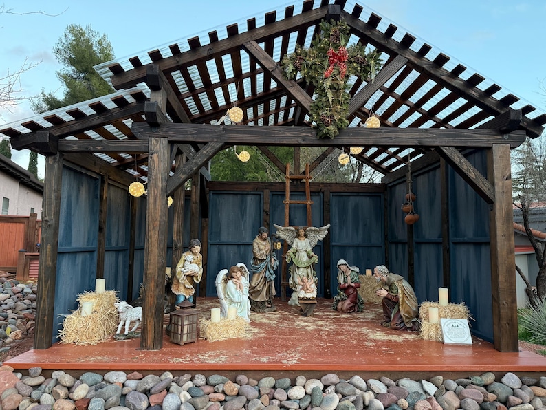 Puede incluir: Un bel&eacute;n navide&ntilde;o con figuras de Mar&iacute;a, Jos&eacute;, &aacute;ngeles y el ni&ntilde;o Jes&uacute;s. La escena est&aacute; ambientada dentro de una estructura de madera con una cruz y luces decorativas. Las figuras est&aacute;n dispuestas sobre una plataforma con balas de heno y velas.