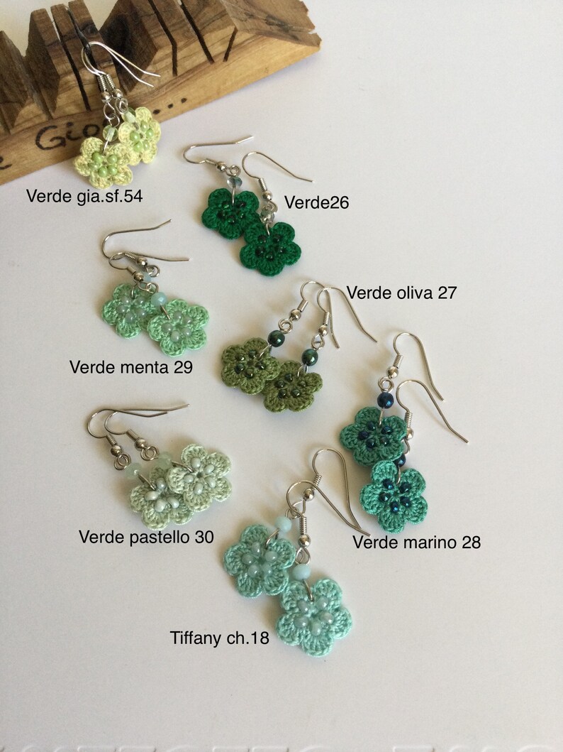 K&ouml;nnte beinhalten: H&auml;kelohrringe in Form von Blumen in verschiedenen Gr&uuml;nt&ouml;nen, darunter Hellgr&uuml;n, Mintgr&uuml;n, Dunkelgr&uuml;n und T&uuml;rkis. Die Ohrringe werden auf einer Holzoberfl&auml;che mit Textbezeichnungen f&uuml;r die Farben angezeigt.