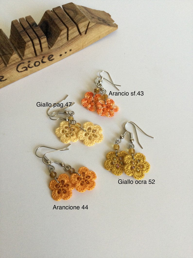 K&ouml;nnte beinhalten: Vier Paare geh&auml;kelter Blumenohrringe in Orange- und Gelbt&ouml;nen. Die Ohrringe sind mit italienischen Namen und Zahlen beschriftet, wie z. B. "Arancio sf.43" und "Giallo ocra 52".