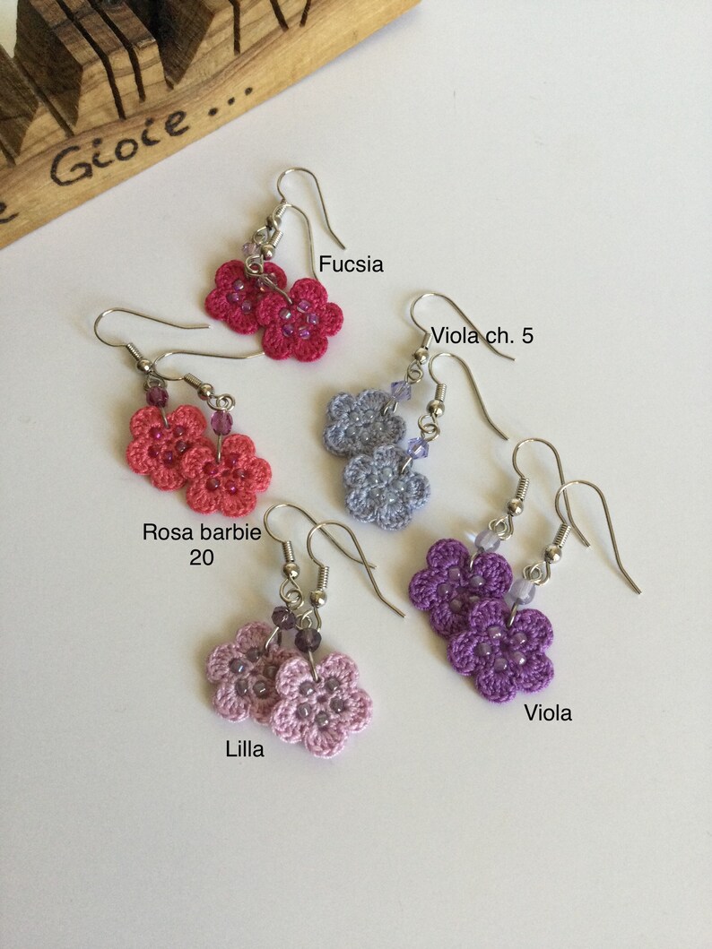 K&ouml;nnte beinhalten: Sechs Paare geh&auml;kelter Blumenohrringe in verschiedenen Farben, darunter Fuchsia, Lila, Rosa und Grau. Jedes Paar besteht aus zwei kleinen geh&auml;kelten Blumen mit einem silbernen Haken.