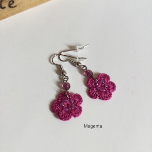 K&ouml;nnte beinhalten: Ein Paar magentafarbene geh&auml;kelte Blumenohrringe mit lila Perlen. Die Ohrringe befinden sich auf einem wei&szlig;en Hintergrund.
