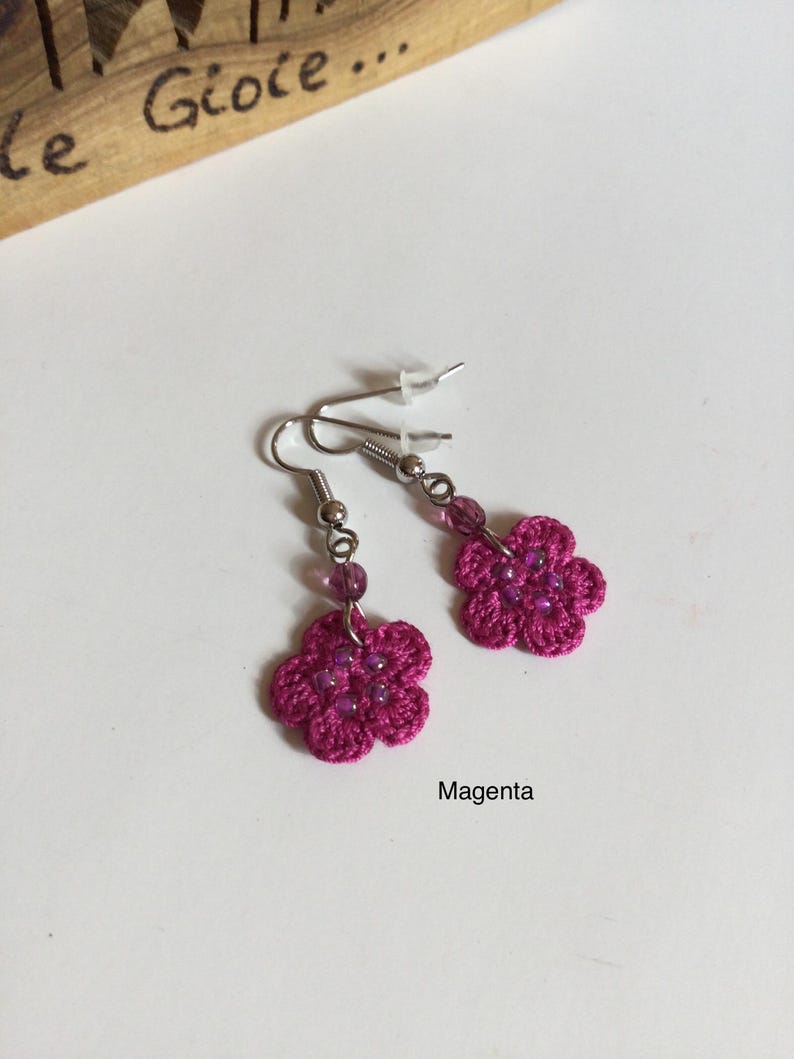 K&ouml;nnte beinhalten: Ein Paar magentafarbene geh&auml;kelte Blumenohrringe mit lila Perlen. Die Ohrringe befinden sich auf einem wei&szlig;en Hintergrund.