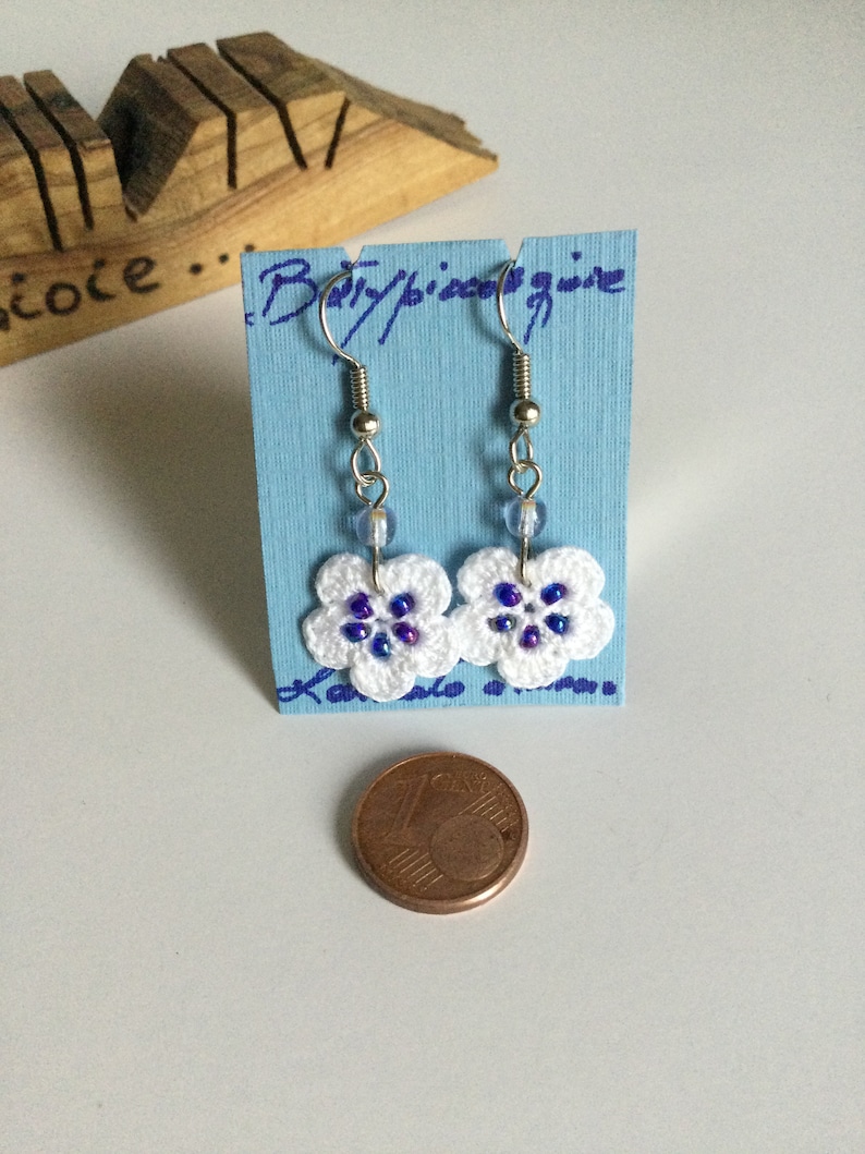 K&ouml;nnte beinhalten: Wei&szlig;e geh&auml;kelte Blumenohrringe mit blauen und violetten Perlen, die an silbernen Haken h&auml;ngen. Die Ohrringe werden auf einer blauen Karte mit dem Text "Baby/piece/wire" pr&auml;sentiert.