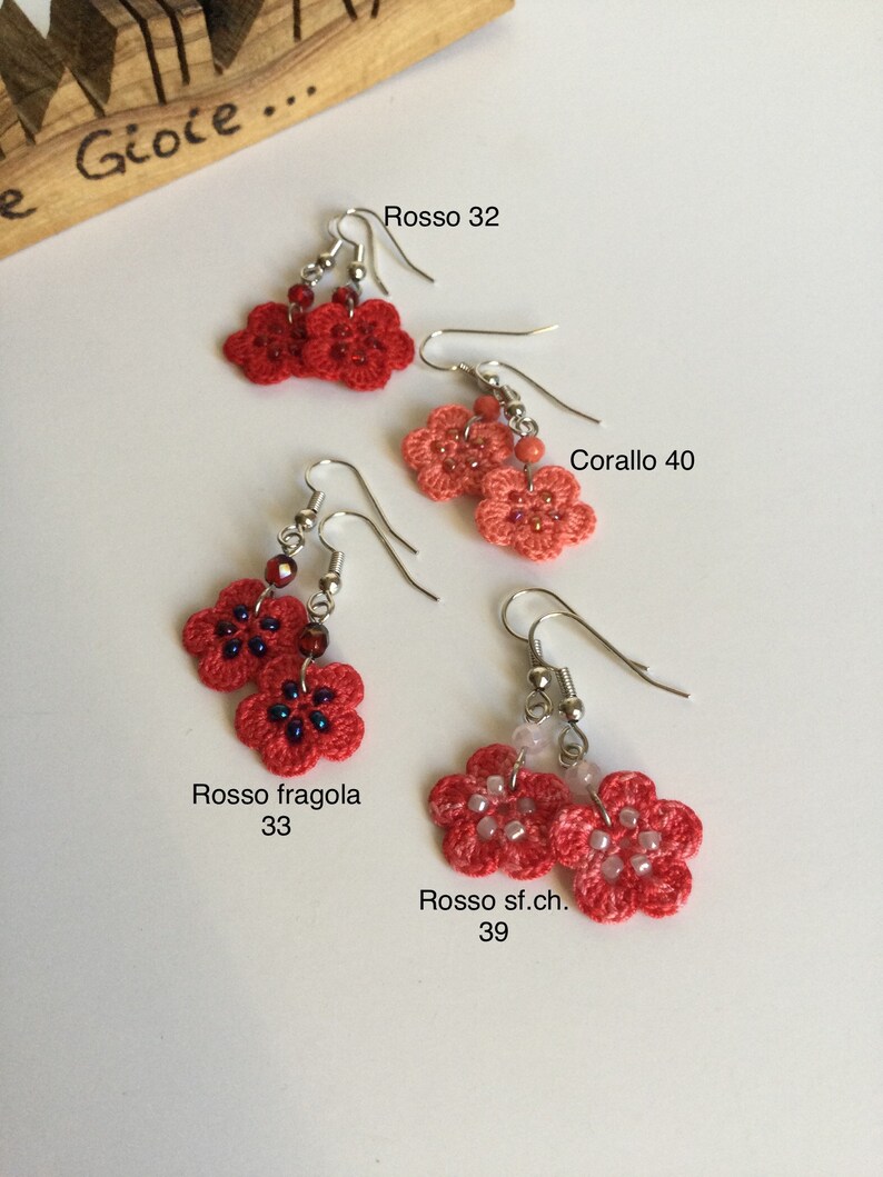 K&ouml;nnte beinhalten: Vier Paare geh&auml;kelter Blumenohrringe in verschiedenen Rott&ouml;nen und Korallenfarben. Die Ohrringe sind mit italienischen Namen und Zahlen beschriftet, wie z. B. "Rosso 32" und "Corallo 40".