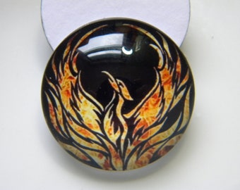 Könnte beinhalten: Ein runder Glascabochon mit schwarzem Hintergrund und einem Phönix-Design in orangefarbenen und gelben Flammen.