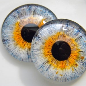 Może przedstawiać: Dwa szklane kaboszony oczu z szczegółowym wzorem tęczówki. Tęczówki mają mieszankę odcieni niebieskiego, białego i pomarańczowego, z ciemną źrenicą pośrodku. Szkło ma błyszczące wykończenie i jest na białym tle.