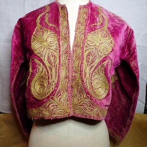 Pu&ograve; includere: Giacca in velluto magenta con ricami dorati elaborati. La giacca presenta un colletto alla coreana e un design frontale aperto e corto. I ricami dorati sono concentrati sui pannelli anteriori e sulle maniche, creando un contrasto.