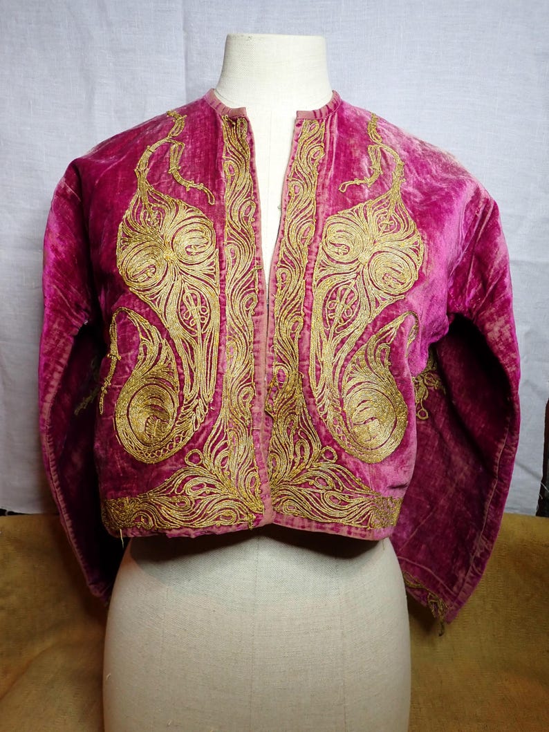 Pu&ograve; includere: Giacca in velluto magenta con ricami dorati elaborati. La giacca presenta un colletto alla coreana e un design frontale aperto e corto. I ricami dorati sono concentrati sui pannelli anteriori e sulle maniche, creando un contrasto.