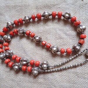 May include: A necklace with coral-coloured beads and silver-toned, decorative beads. The necklace has a double strand with a geometric pendant. The jewellery is displayed on a textured, neutral-toned surface.