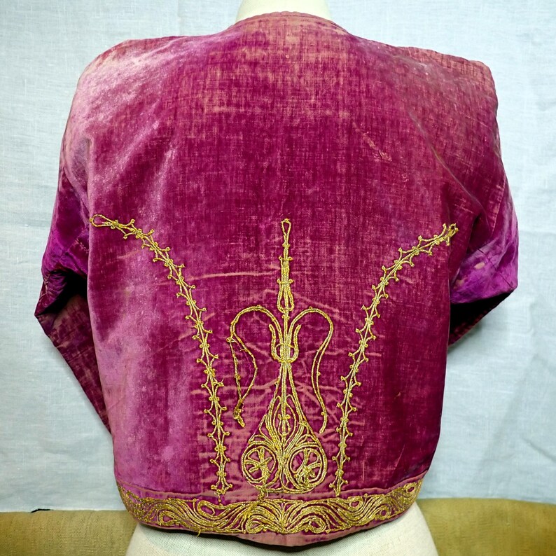 Pu&ograve; includere: Una giacca in velluto magenta con ricami dorati. La giacca presenta ricami dorati ornati sul retro, con un design decorativo e un bordo dettagliato lungo il fondo. Le maniche sono corte e la giacca &egrave; esposta su un manichino.