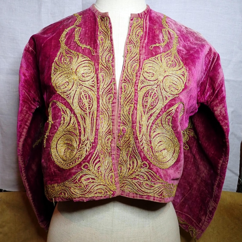 Pu&ograve; includere: Giacca in velluto magenta con ricami dorati. La giacca presenta un davanti aperto e intricati disegni in filo d'oro sui pannelli anteriori e sulle maniche. Il ricamo ha un motivo a forma di paisley. La giacca &egrave; esposta su un manichino.