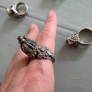 Può includere: Primo piano di tre anelli d'argento. L'anello principale presenta un design ornato a forma di gabbia con un gruppo di piccole sfere sulla parte superiore. Gli altri anelli hanno motivi dettagliati. Gli anelli sono su una superficie azzurra.