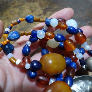 May include: A multi-strand necklace featuring a variety of beads in shades of blue, brown, orange, and white. The necklace includes large amber-colored beads and a silver-colored metal clasp. The beads are strung together to create a unique and colorful accessory.