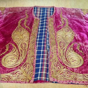 Pu&ograve; includere: Giacca in velluto magenta con ricami dorati. La giacca presenta una fodera a quadri blu e bianca e intricati motivi dorati sul davanti e sulle maniche. L'indumento &egrave; steso su una superficie marrone chiaro.