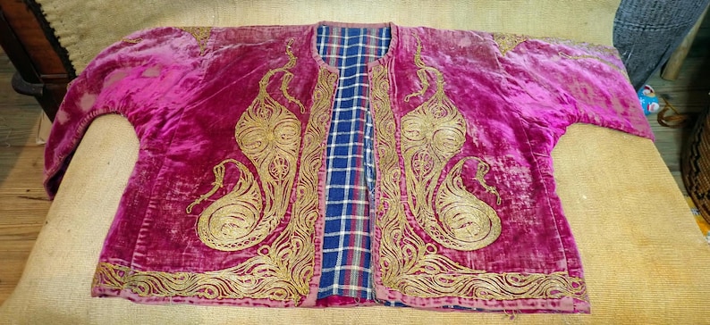 Pu&ograve; includere: Giacca in velluto magenta con ricami dorati. La giacca presenta una fodera a quadri blu e bianca e intricati motivi dorati sul davanti e sulle maniche. L'indumento &egrave; steso su una superficie marrone chiaro.
