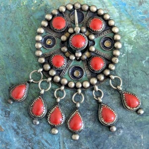 May include: A circular silver brooch featuring a central red stone surrounded by five smaller red stones and decorative metal beading. Six teardrop-shaped red stones hang from the bottom edge of the brooch, each attached by a small silver ring.