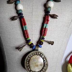 May include: A multi-strand necklace featuring red, turquoise, and white beads, accented with metal charms and a large pendant. The pendant has a silver-colored frame and a light-colored oval center. The necklace is displayed on a mannequin.